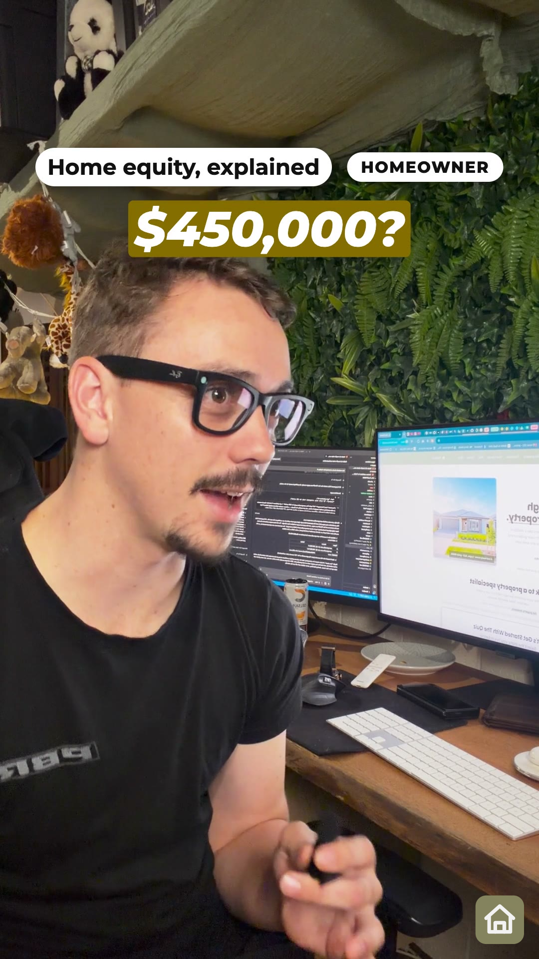 A man in glasses and a black T-shirt talking at a desk with two monitors and a green plant wall behind