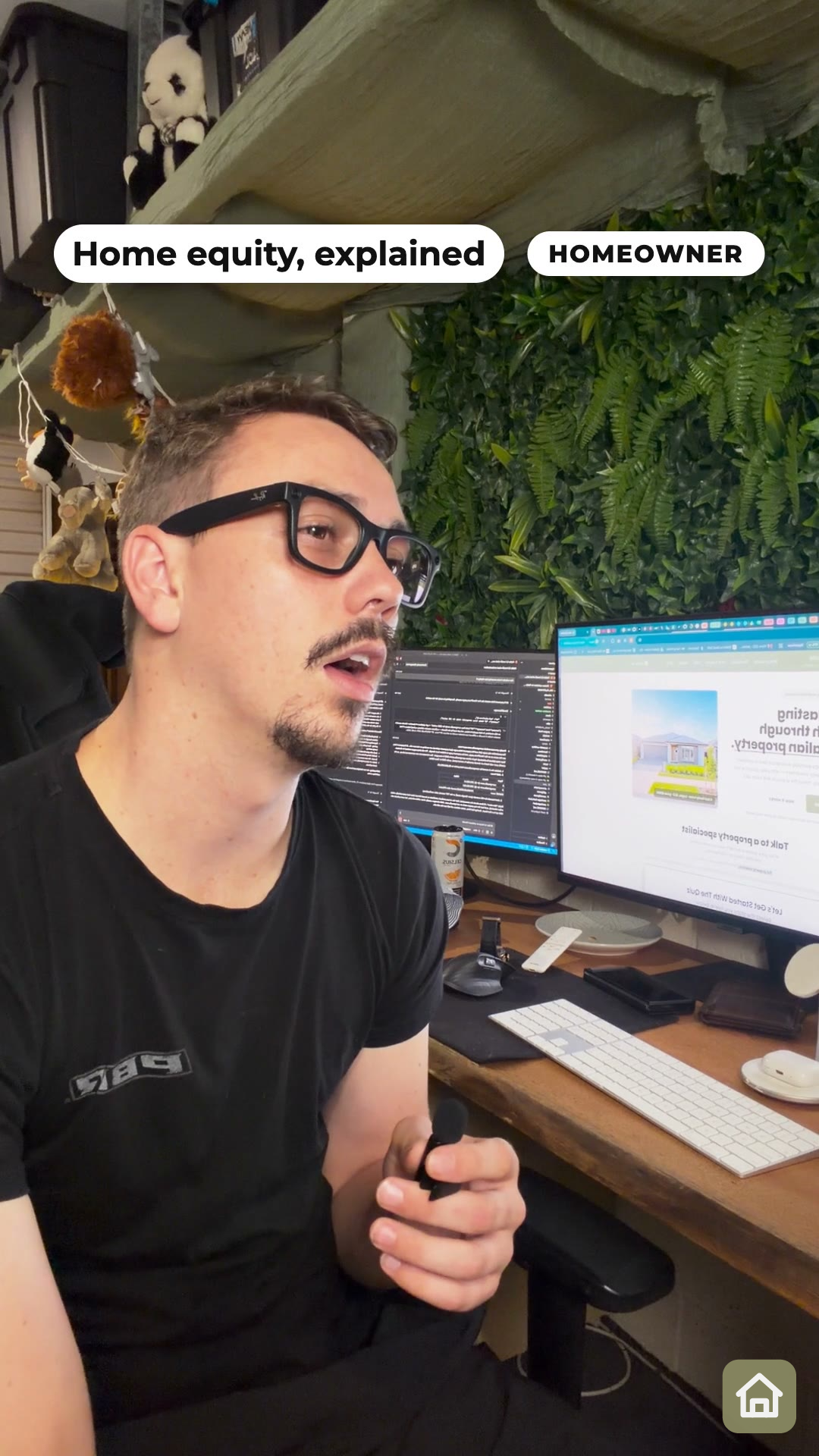 The same man holding a small microphone at his desk, with computer screens and leafy greenery behind him